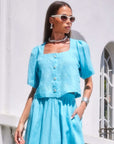 Woman wearing a sky blue Pia Poof Sleeve Linen Top with square neck and button front closure paired with matching high waist skirt