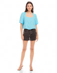 Woman wearing light blue Pia Poof Sleeve Linen Top with square neck and button front paired with black denim shorts