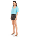 Woman wearing a light blue Pia Poof Sleeve Linen Top with square neck and button front closure paired with black shorts and heels