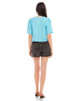 Woman wearing a light blue Pia Poof Sleeve Linen Top with short puff sleeves, paired with black denim shorts, shown from the back