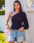 Woman wearing a black Talia One-Shoulder Top with elasticized waist and one long blouson sleeve paired with ripped denim shorts