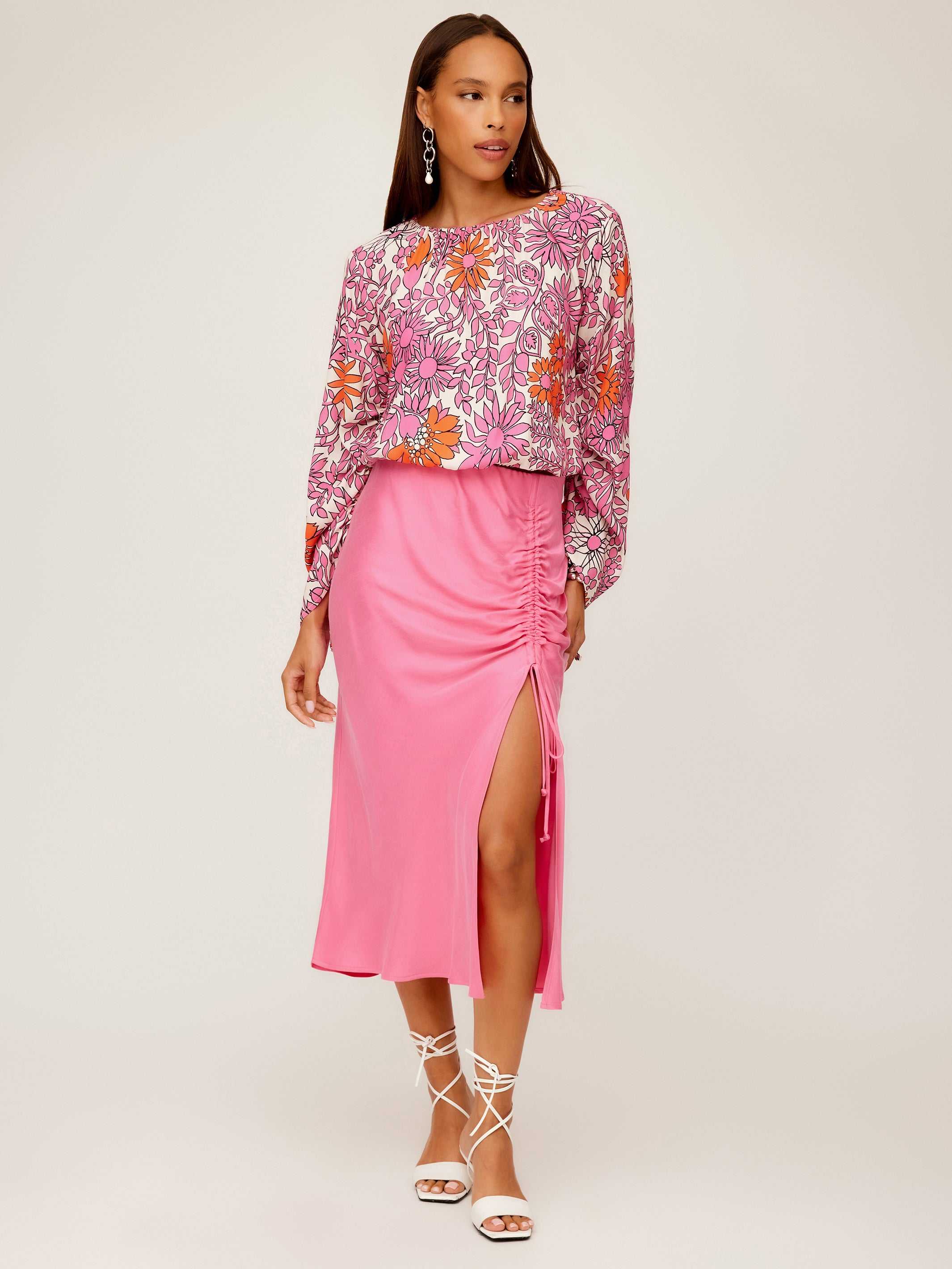 Woman wearing a pink Fiona Shirred Midi Skirt with side shirring and high slit paired with a floral long-sleeve blouse and white lace-up sandals
