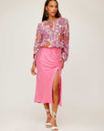 Woman wearing a pink Fiona Shirred Midi Skirt with side shirring and high slit paired with a floral long-sleeve blouse and white lace-up sandals