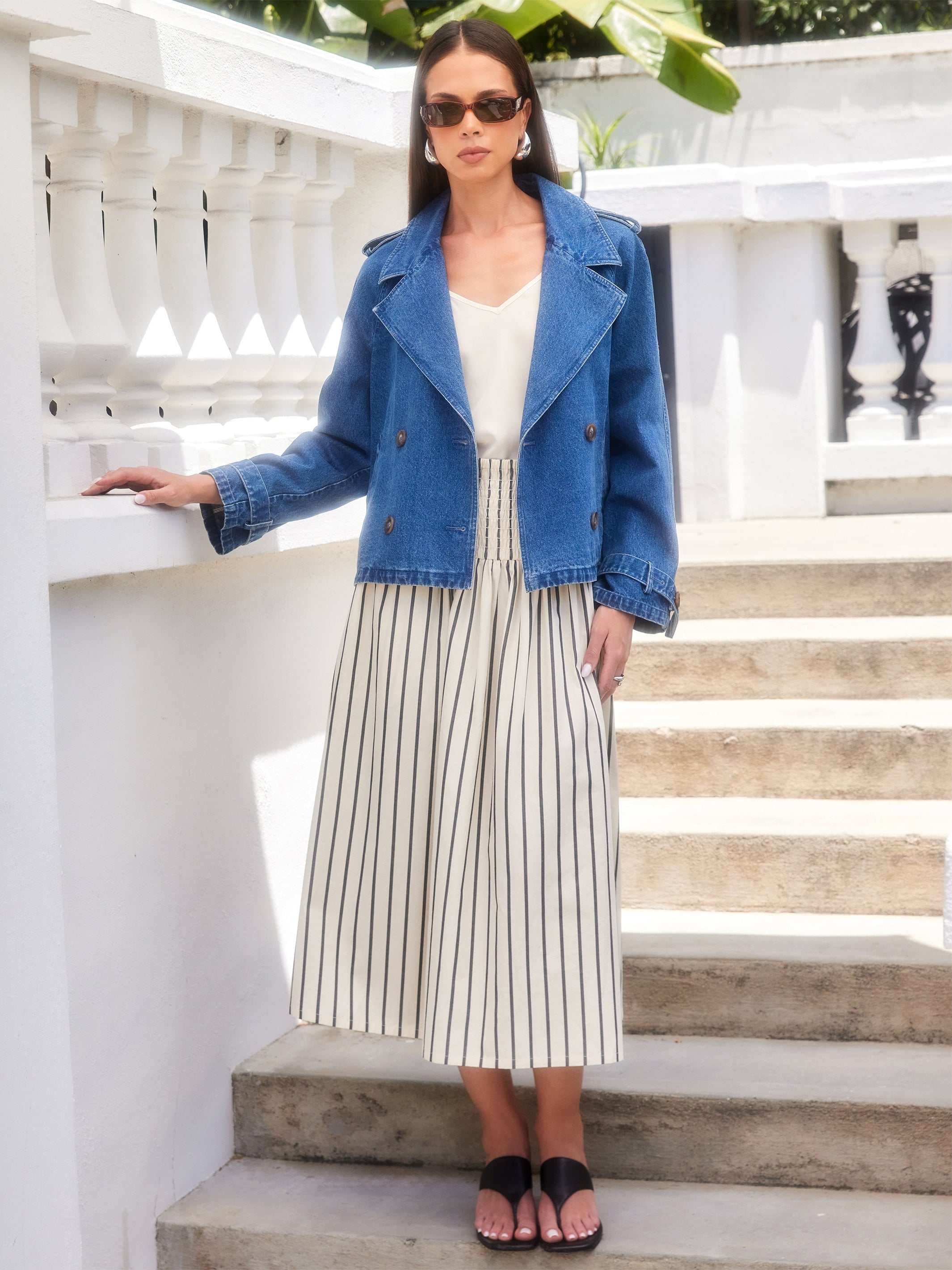 Woman wearing a Chelsea Cropped Trench denim jacket with double breast buttons and notch collar over a white striped dress