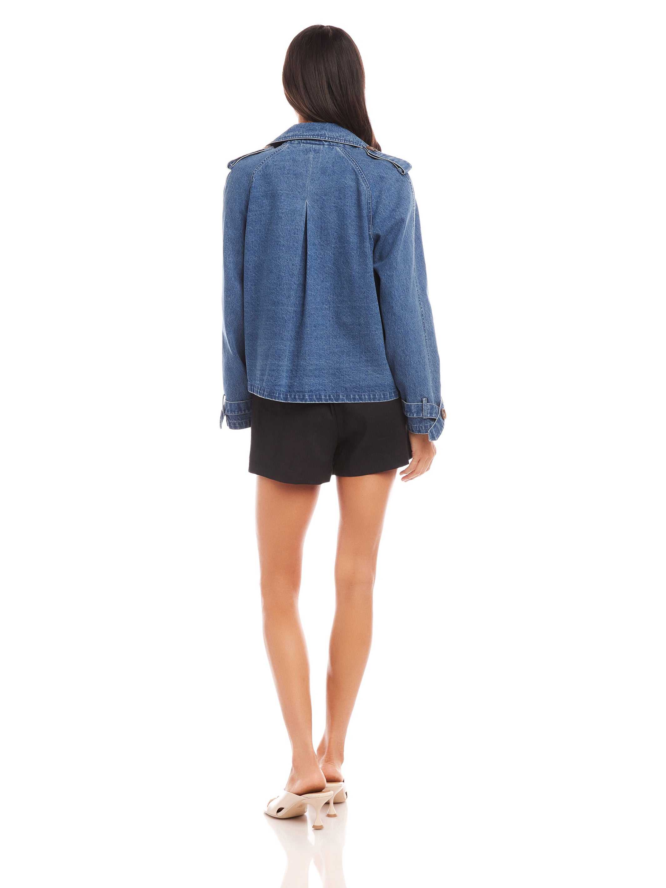 Woman wearing a blue denim Chelsea Cropped Trench jacket with long raglan sleeves and boxy fit shown from the back
