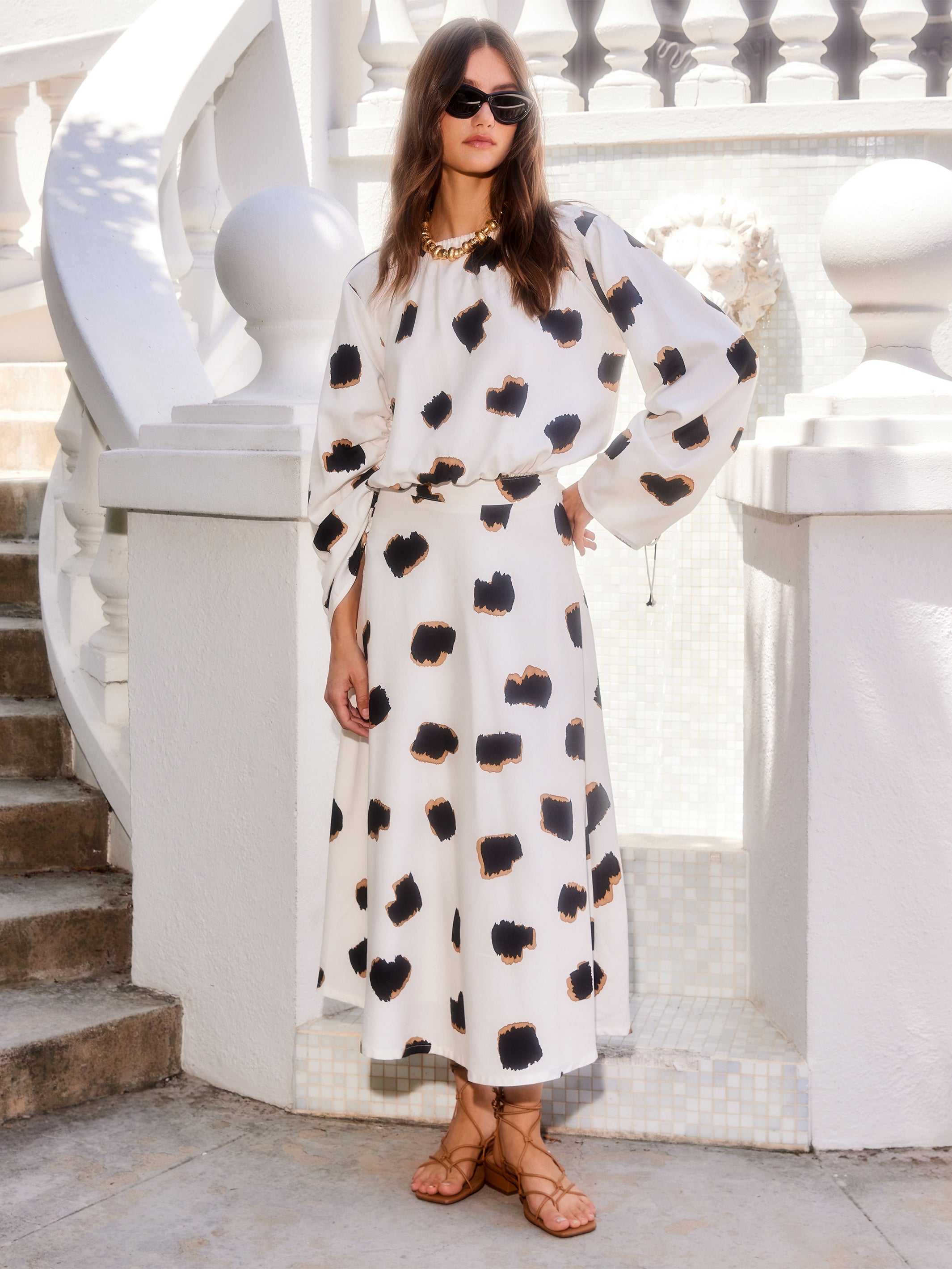 Woman wearing a high-waisted, flowy Lexi Skirt midi in white Sahara Twill print with black and tan spots, paired with a matching top and sandals.
