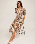 Woman wearing a brown and white paisley print Poppy Shirred Waist Dress with puff sleeves and side slit sitting on a stool