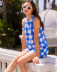 Woman wearing blue and white checkered sleeveless dress sitting outdoors on a white ledge with palm trees in the background