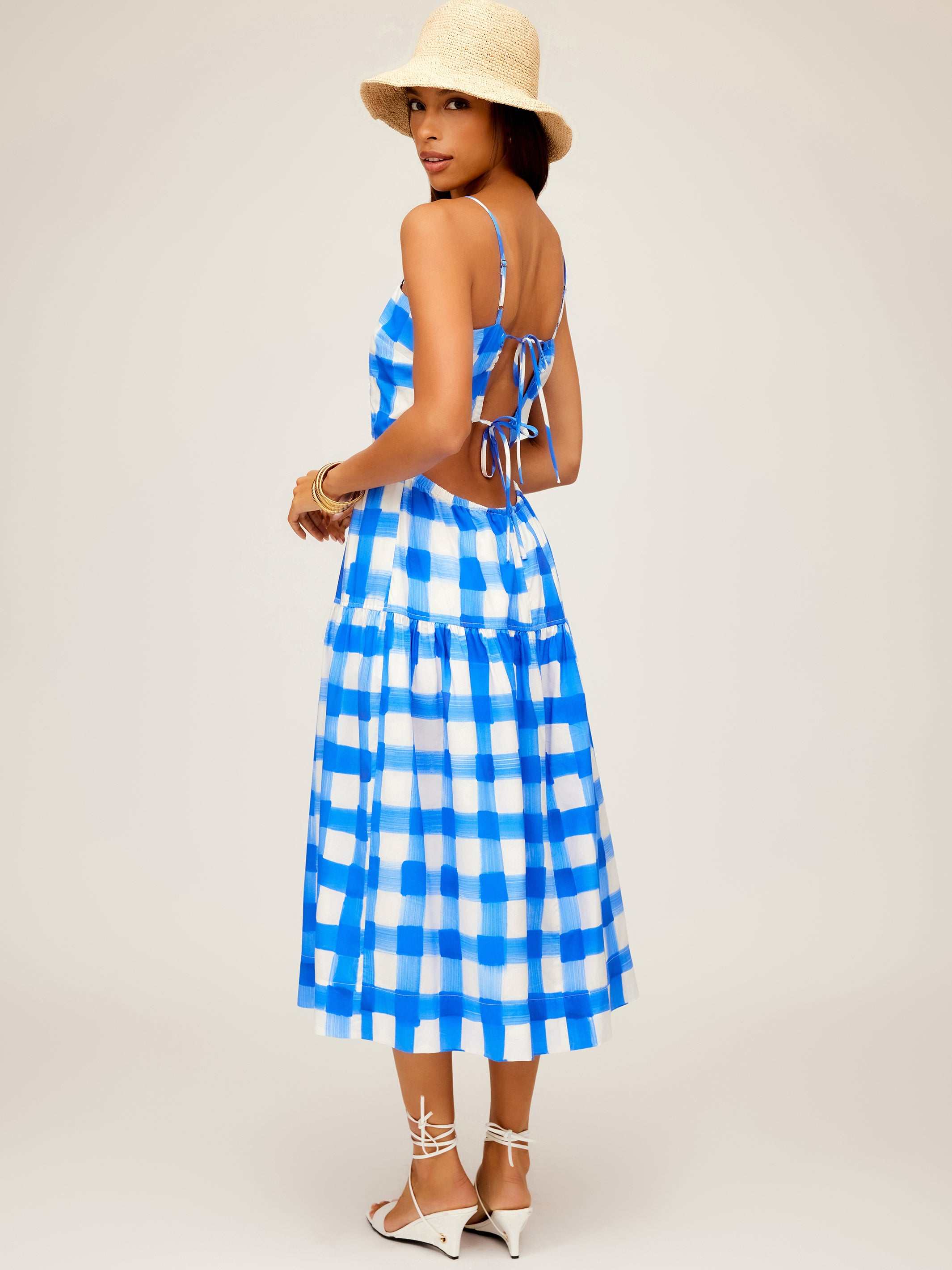 Woman wearing blue and white checkered Costa Dress with open back and adjustable straps, paired with straw hat and white sandals