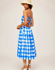 Woman wearing blue and white checkered Costa Dress with open back and adjustable straps, paired with straw hat and white sandals