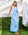 Woman wearing a blue painted paisley print Sera A-Line Dress with adjustable spaghetti straps and maxi length standing outdoors near white stairs and umbrellas