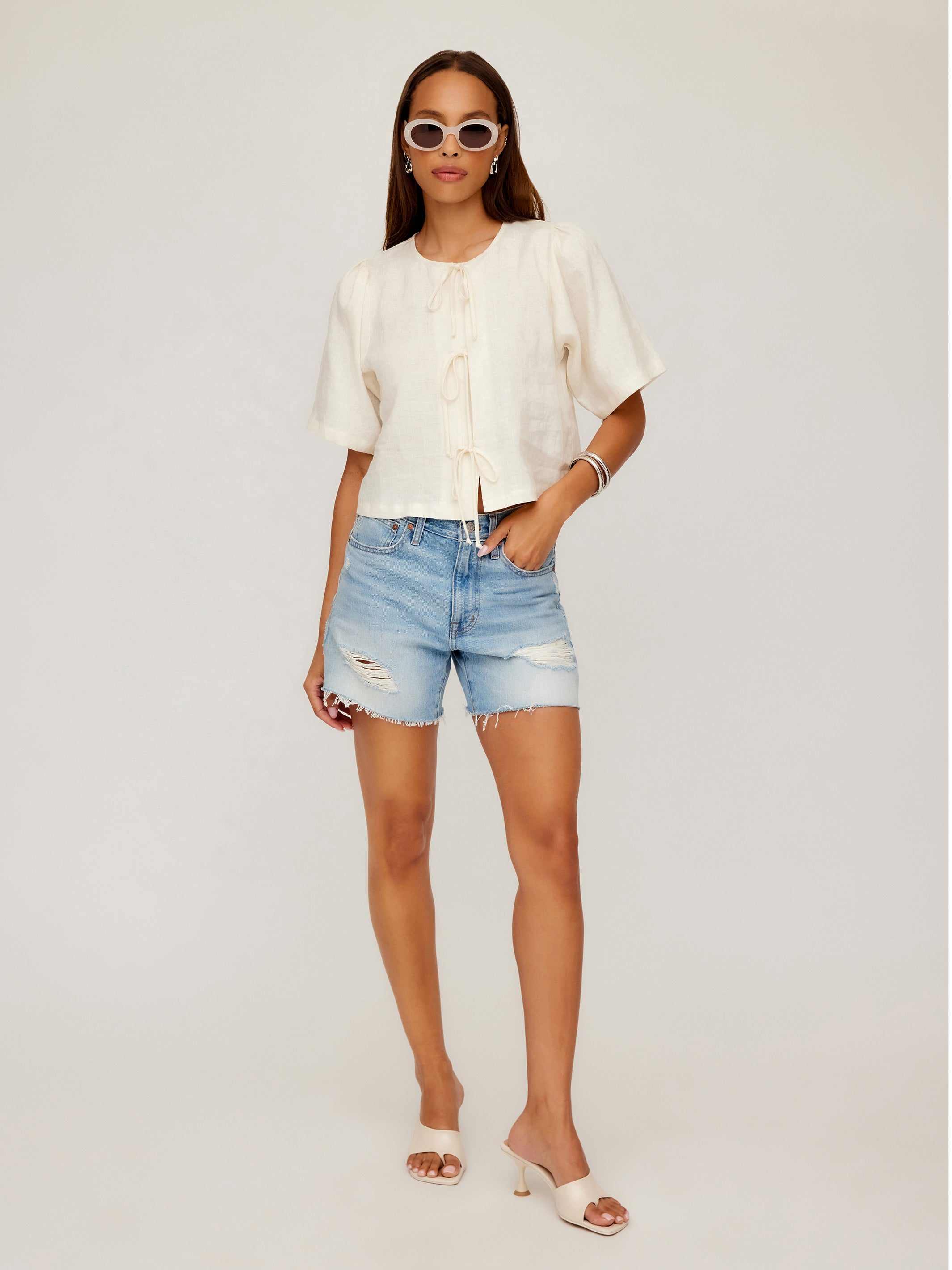 Woman wearing Nadine Bow Detail Top with front tie closure and short puff sleeves paired with distressed high waist denim shorts