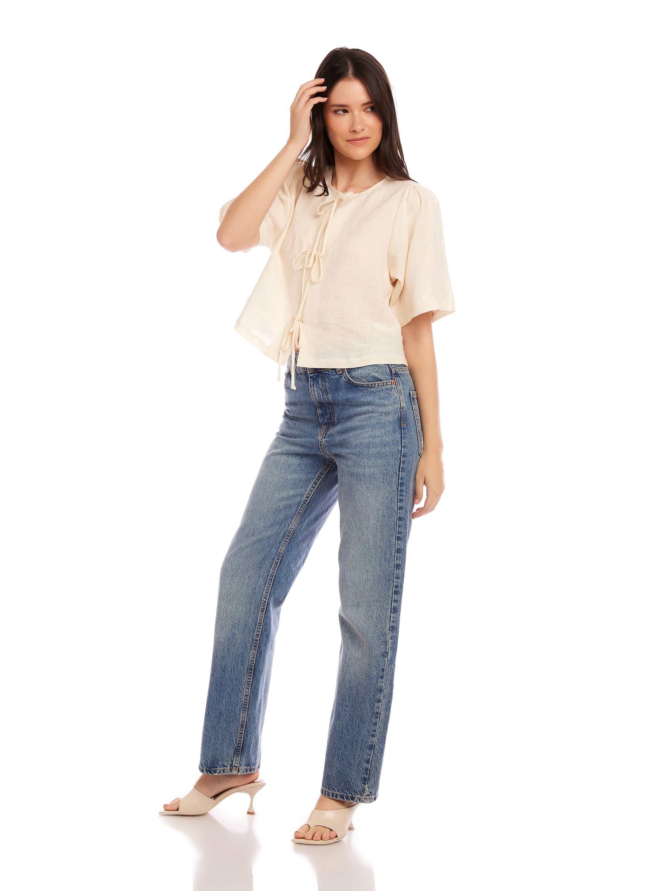 Woman wearing Nadine Bow Detail top in cream linen with front tie and short puff sleeves, paired with high-waisted blue jeans and heels