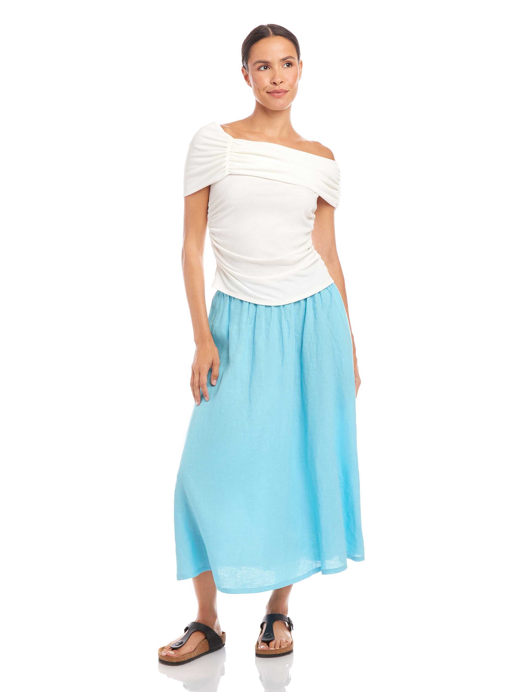 Woman wearing a white Lizzie One-Shoulder Top with asymmetrical sleeves and side shirring, paired with a light blue skirt and black sandals.