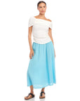 Woman wearing a white Lizzie One-Shoulder Top with asymmetrical sleeves and side shirring, paired with a light blue skirt and black sandals.