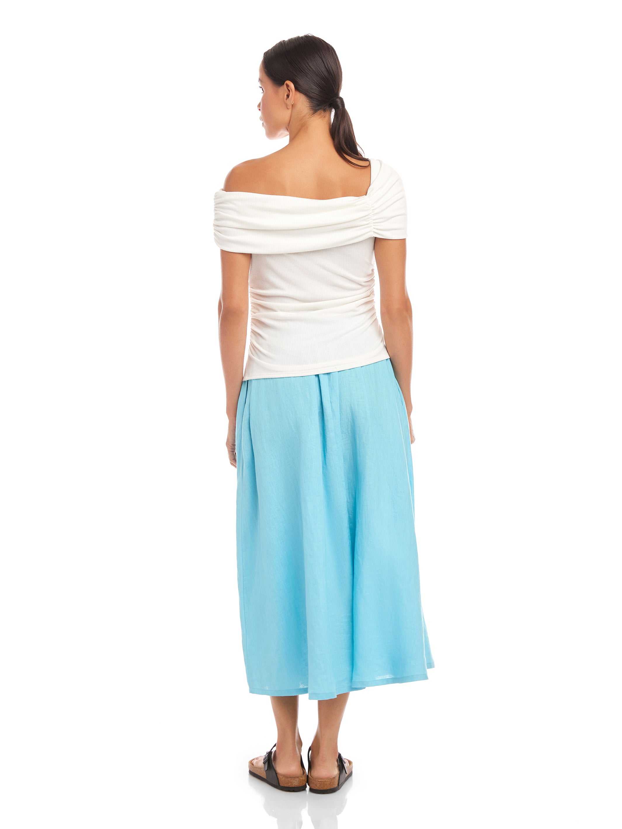 Woman wearing a white Lizzie One-Shoulder Top with asymmetrical sleeves and side shirring, paired with light blue wide-leg pants.