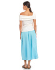 Woman wearing a white Lizzie One-Shoulder Top with asymmetrical sleeves and side shirring, paired with light blue wide-leg pants.