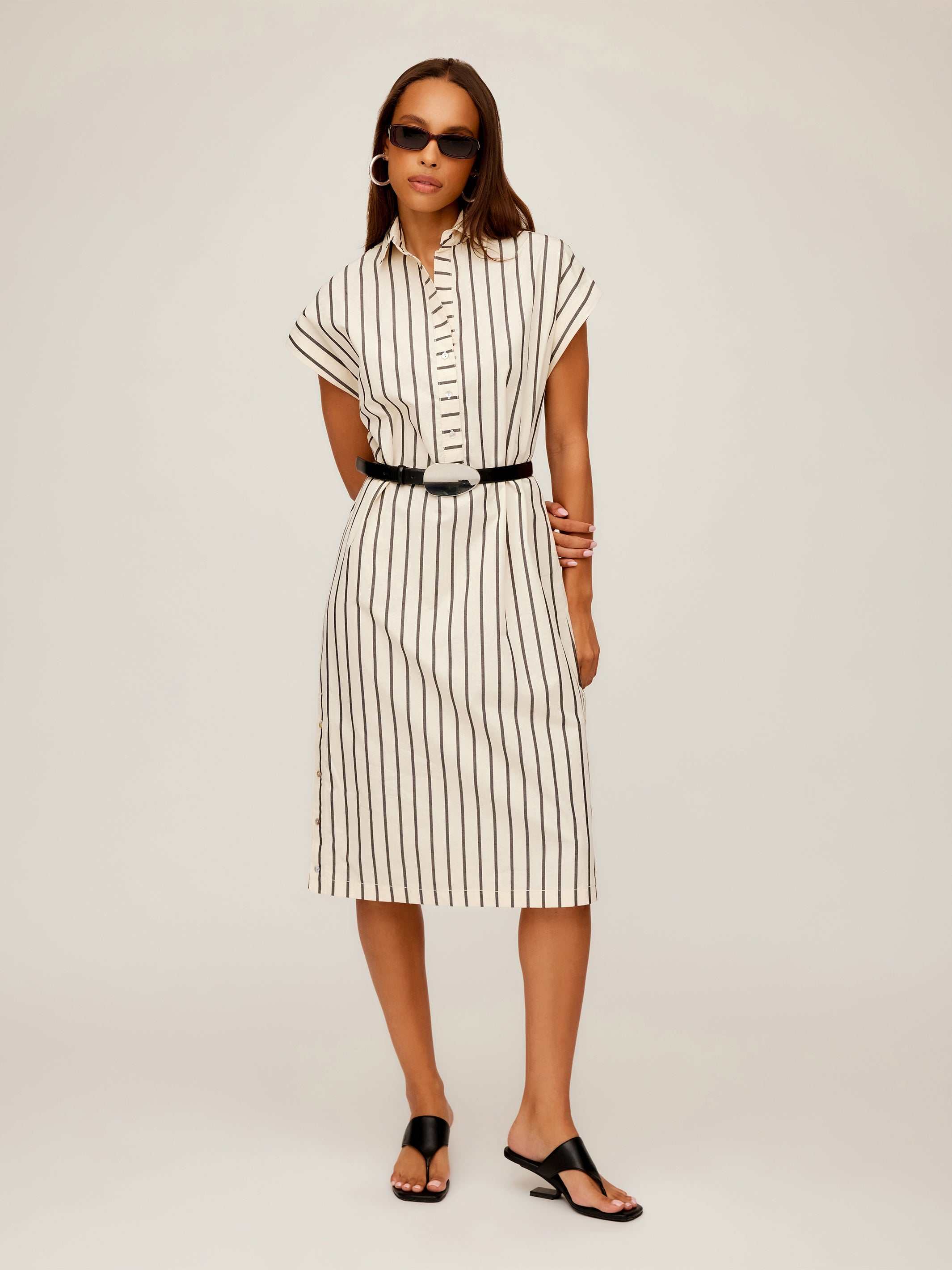 Woman wearing a collared Lucia Stripe Dress with vertical black stripes, cap sleeves, a boxy silhouette, side slit, and black belt at the waist