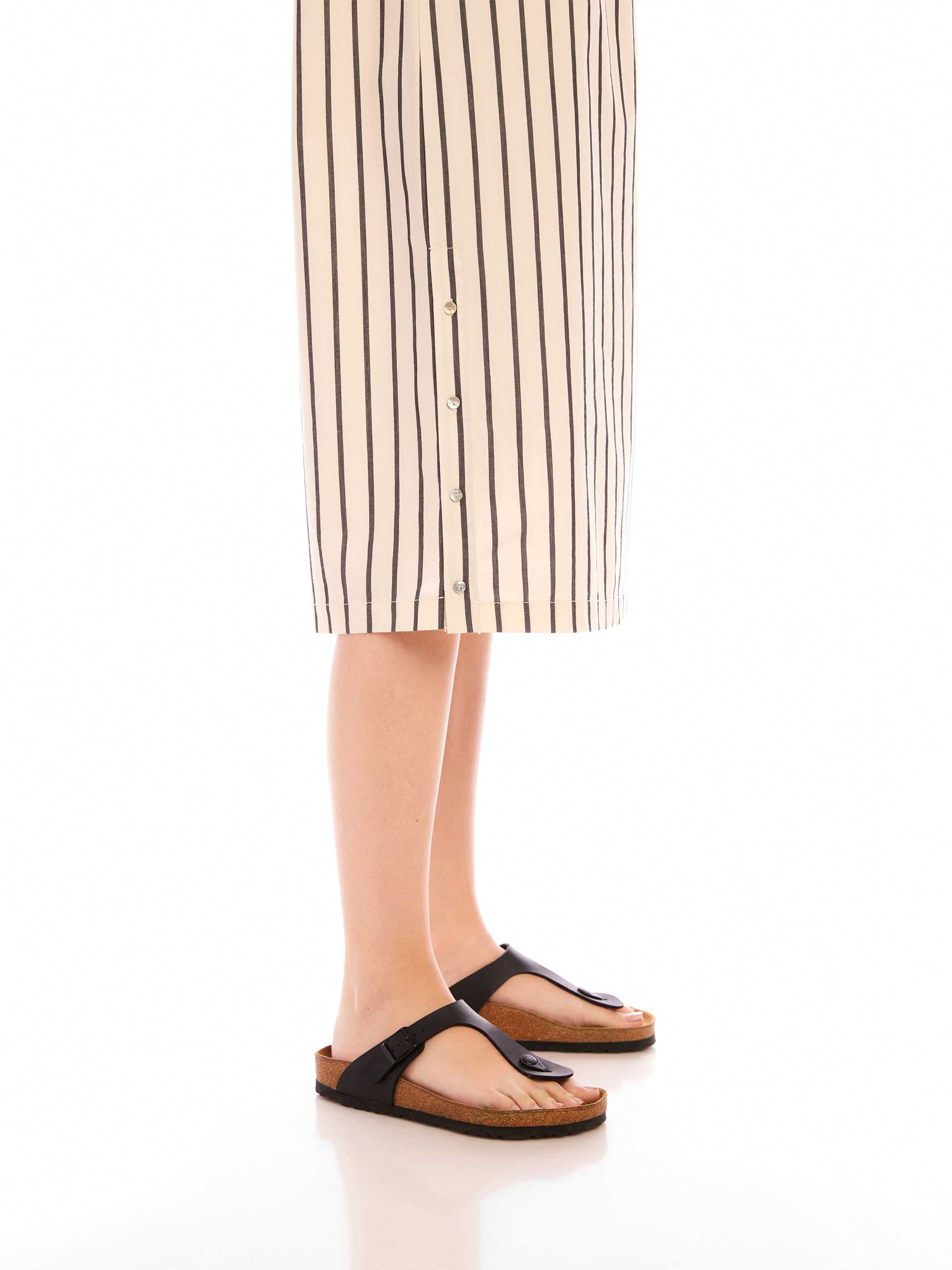Lucia Stripe Dress beige cotton with black vertical stripes, button detail at side slit, worn with black sandals.