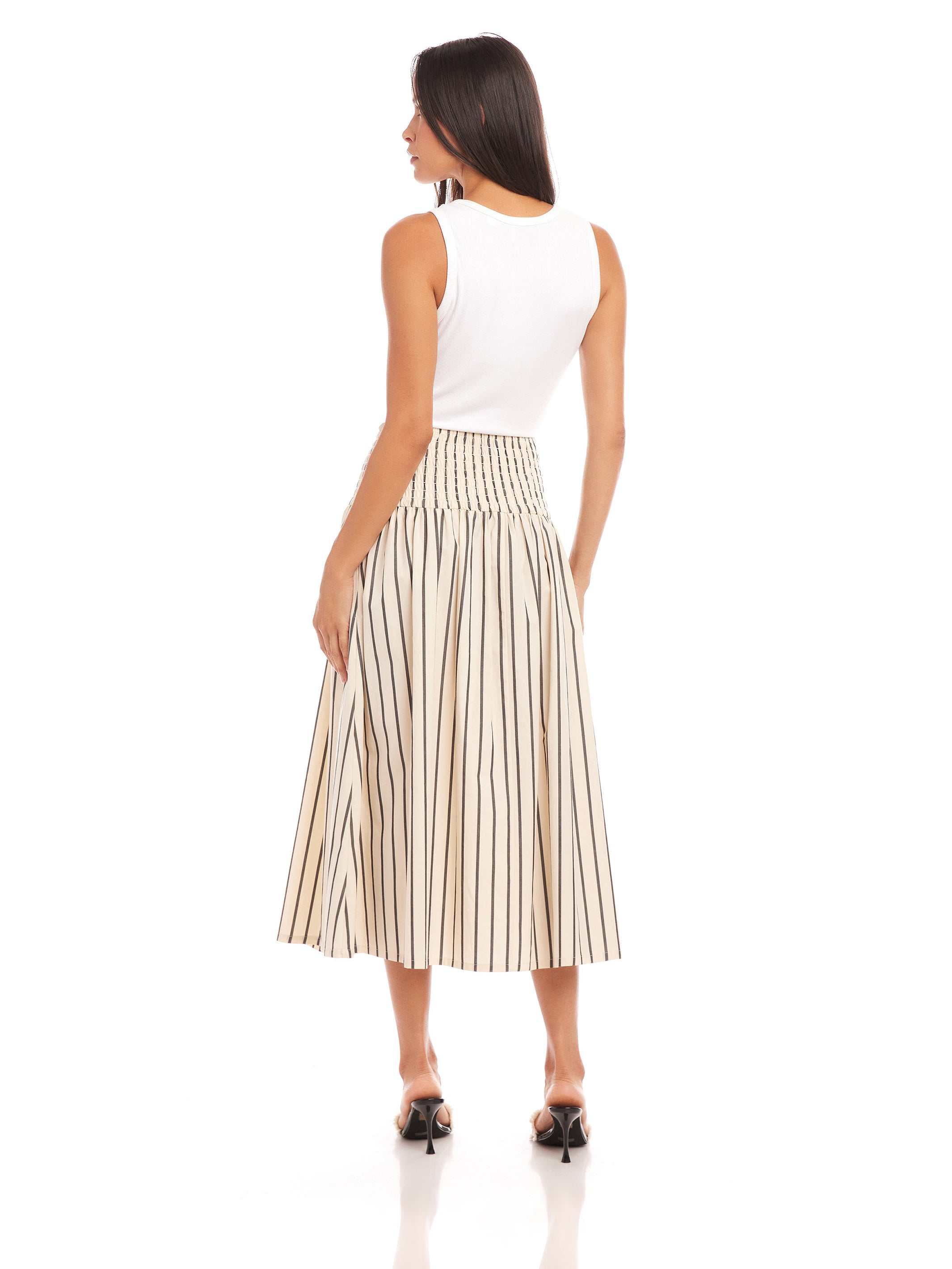 Woman wearing a Rhea Stripe Skirt midi length striped cotton skirt with elasticized sheered waist, styled with a white sleeveless top and black heels.