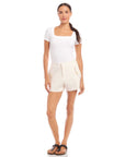 Woman wearing ecru Alma shorts with stretch fabric, side pockets, and pearl buttons, styled with a white top and sandals.