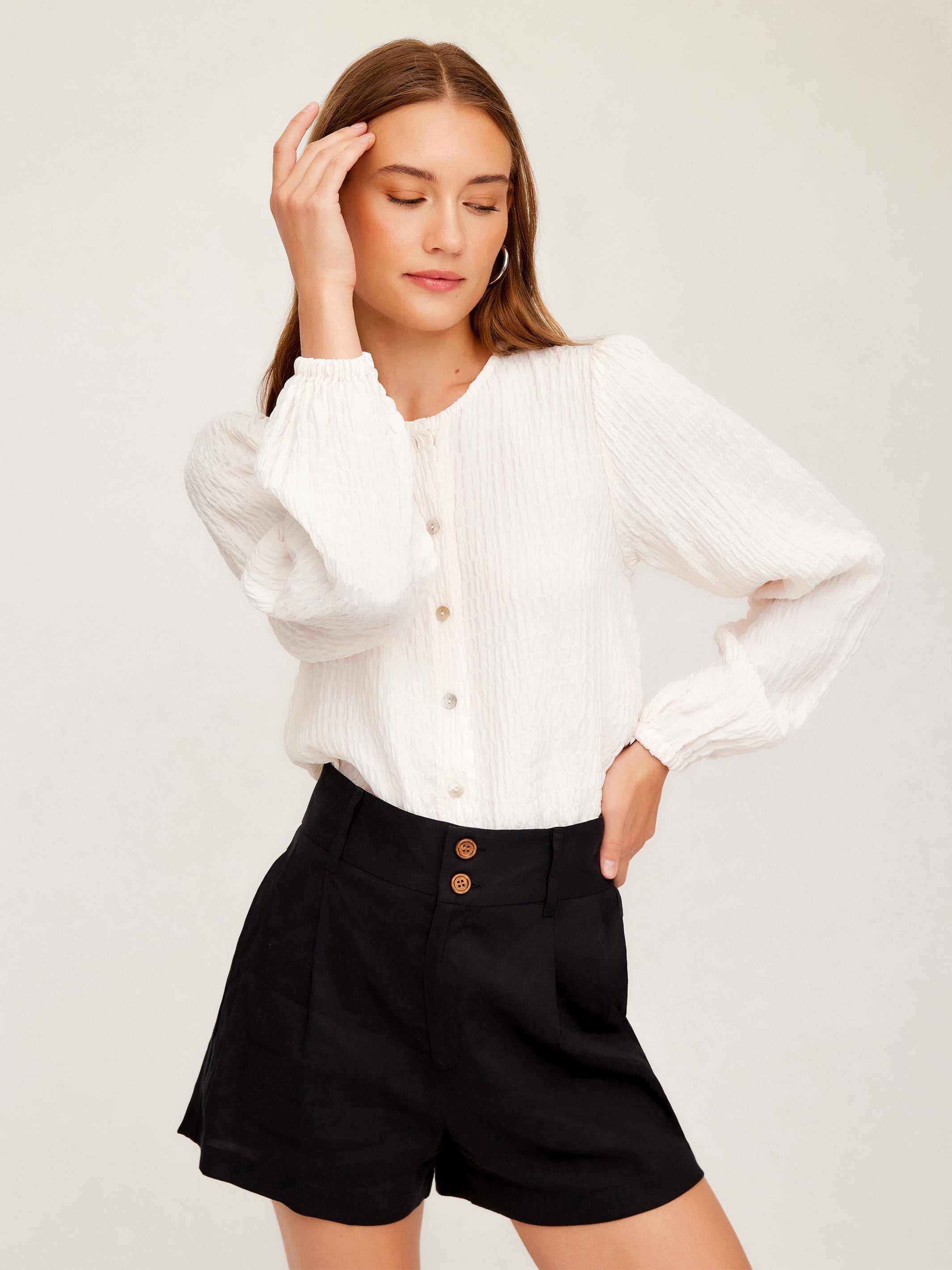 Woman wearing white Aubrey Button-Up Blouse with long bishop sleeves and round neck, paired with black high-waisted shorts.
