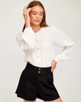 Woman wearing white Aubrey Button-Up Blouse with long bishop sleeves and round neck, paired with black high-waisted shorts.