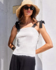 Woman wearing a fitted white Annie Ribbon Tie Top with black adjustable ribbon straps, paired with black pants and a straw hat.