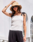 Woman wearing white Annie Ribbon Tie Top with black tie straps and black pants, styled with straw hat and sunglasses outdoors