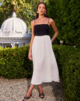 Model wearing Anetta Contrast Midi Dress with fitted black bodice, flowy white skirt, adjustable spaghetti straps, and square neck in outdoor garden setting