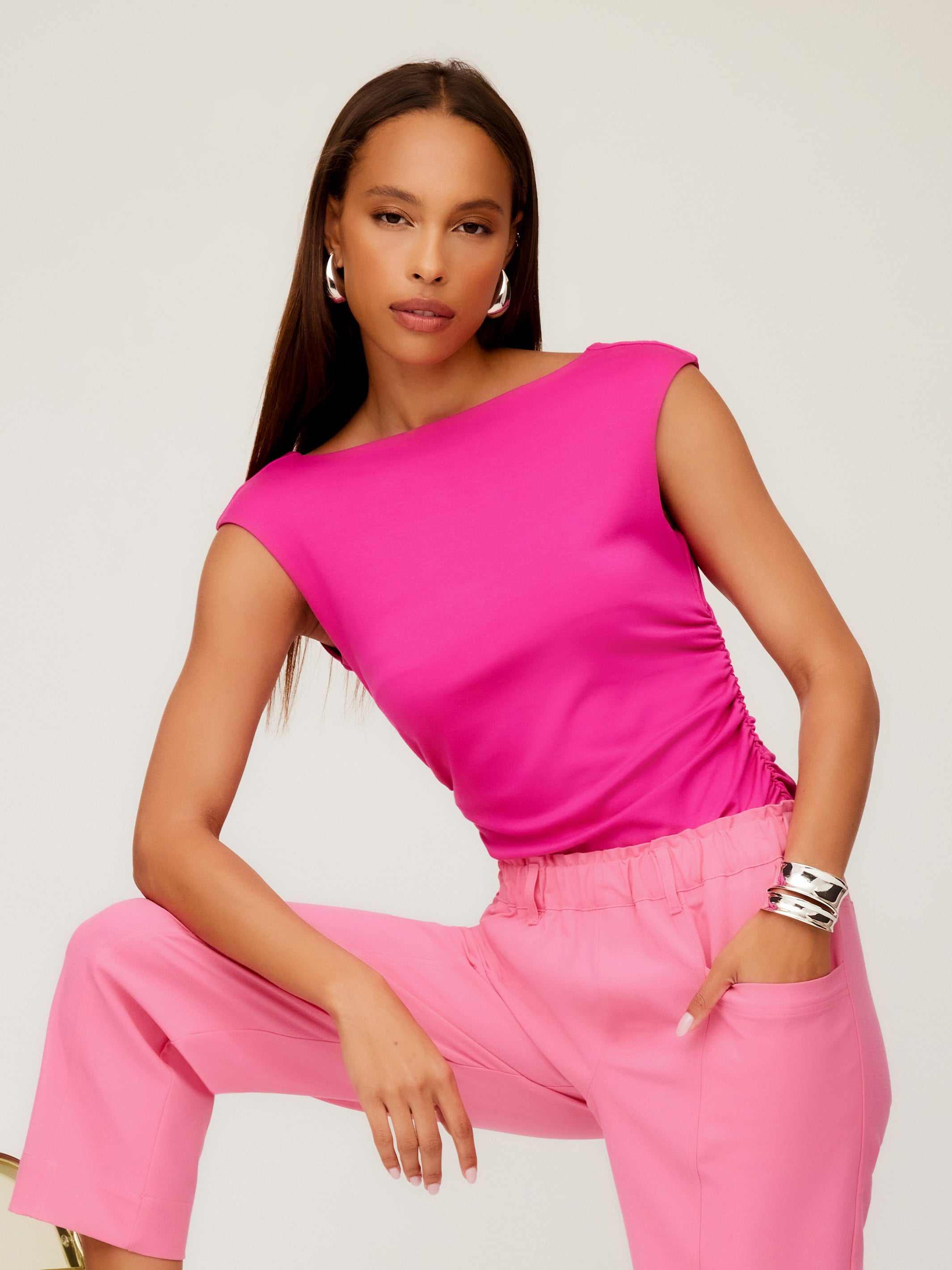Woman wearing vibrant pink Tasha Side-Shirred Top with cap sleeves and boatneck paired with light pink pants.
