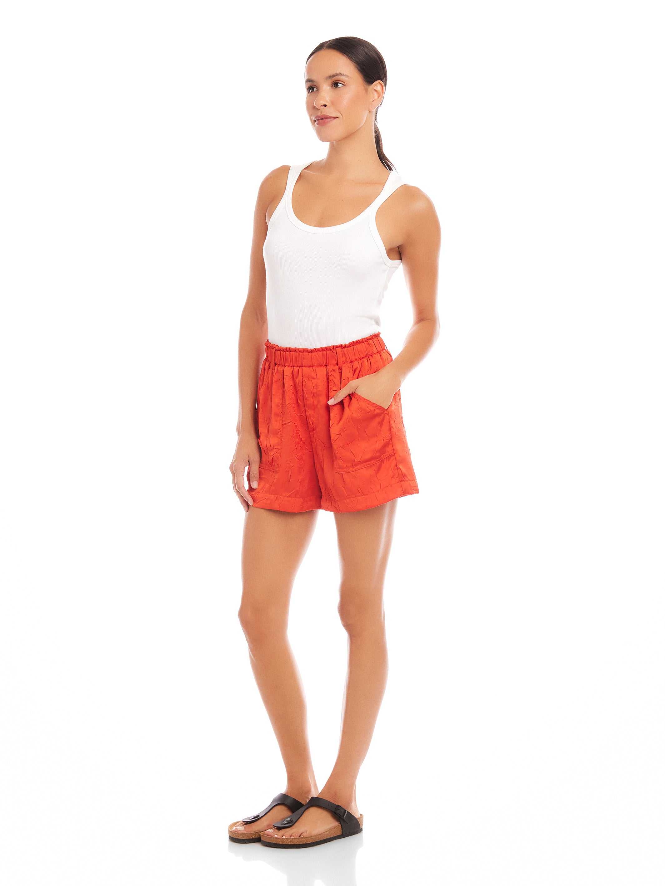 Woman wearing high-waist orange Becca Shorts with elasticized waist and front pockets styled with a white tank top and black sandals