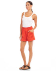 Woman wearing high-waist orange Becca Shorts with elasticized waist and front pockets styled with a white tank top and black sandals