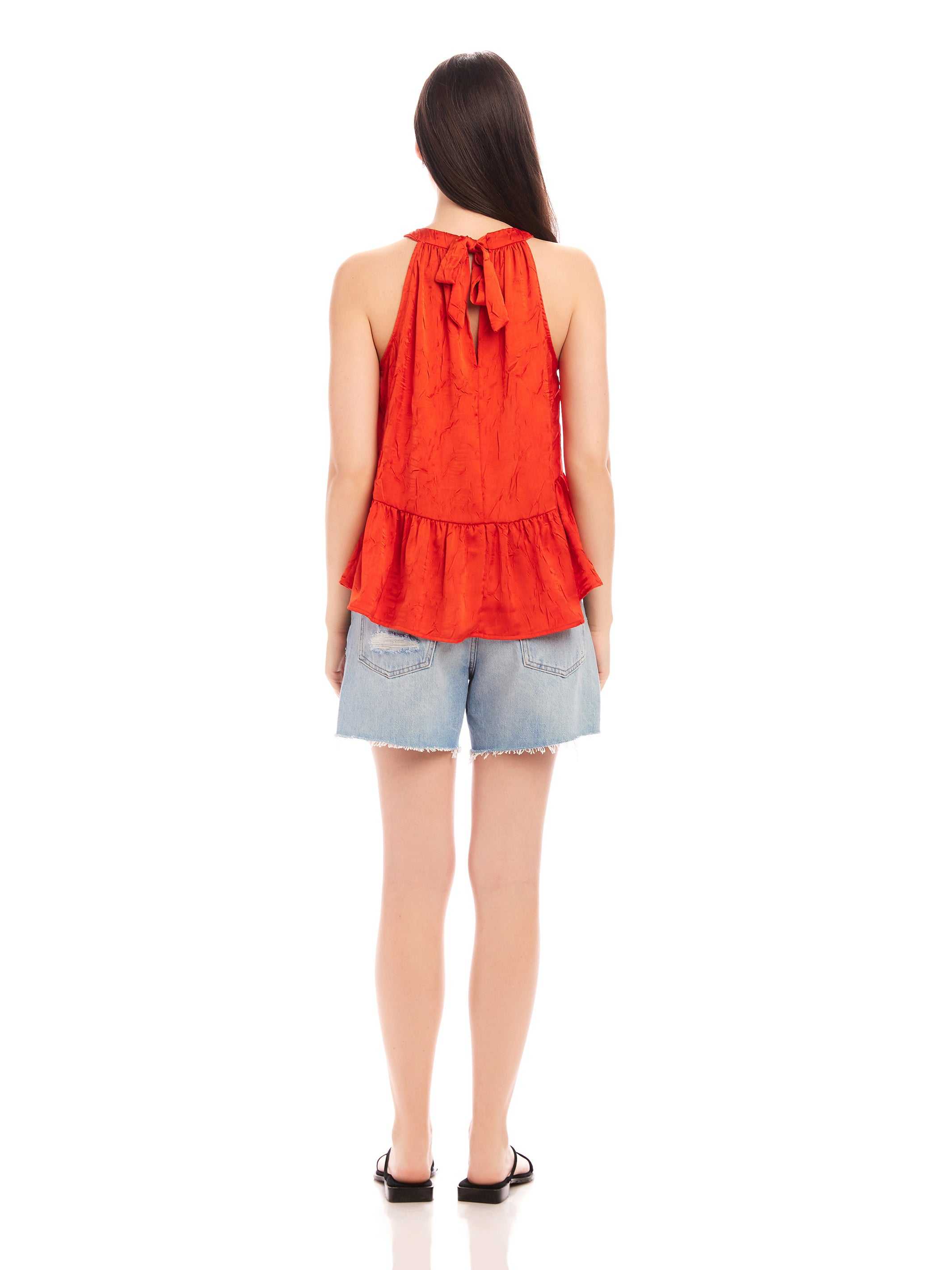 Woman wearing red Floren Ruffle Hem halter top with back tie and denim shorts, back view
