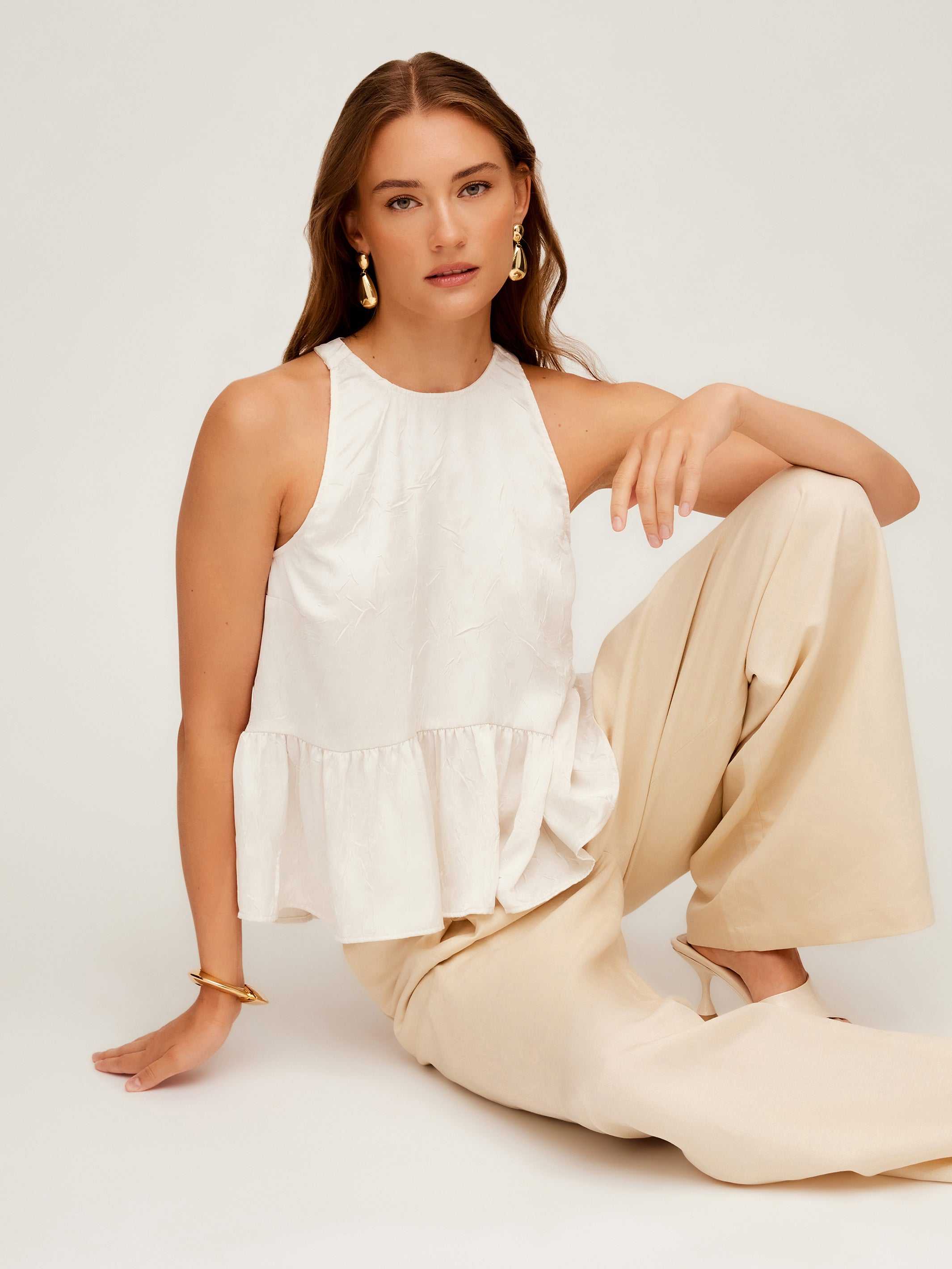 Woman wearing white Floren Ruffle Hem halter top with high-low hem and beige pants sitting on floor