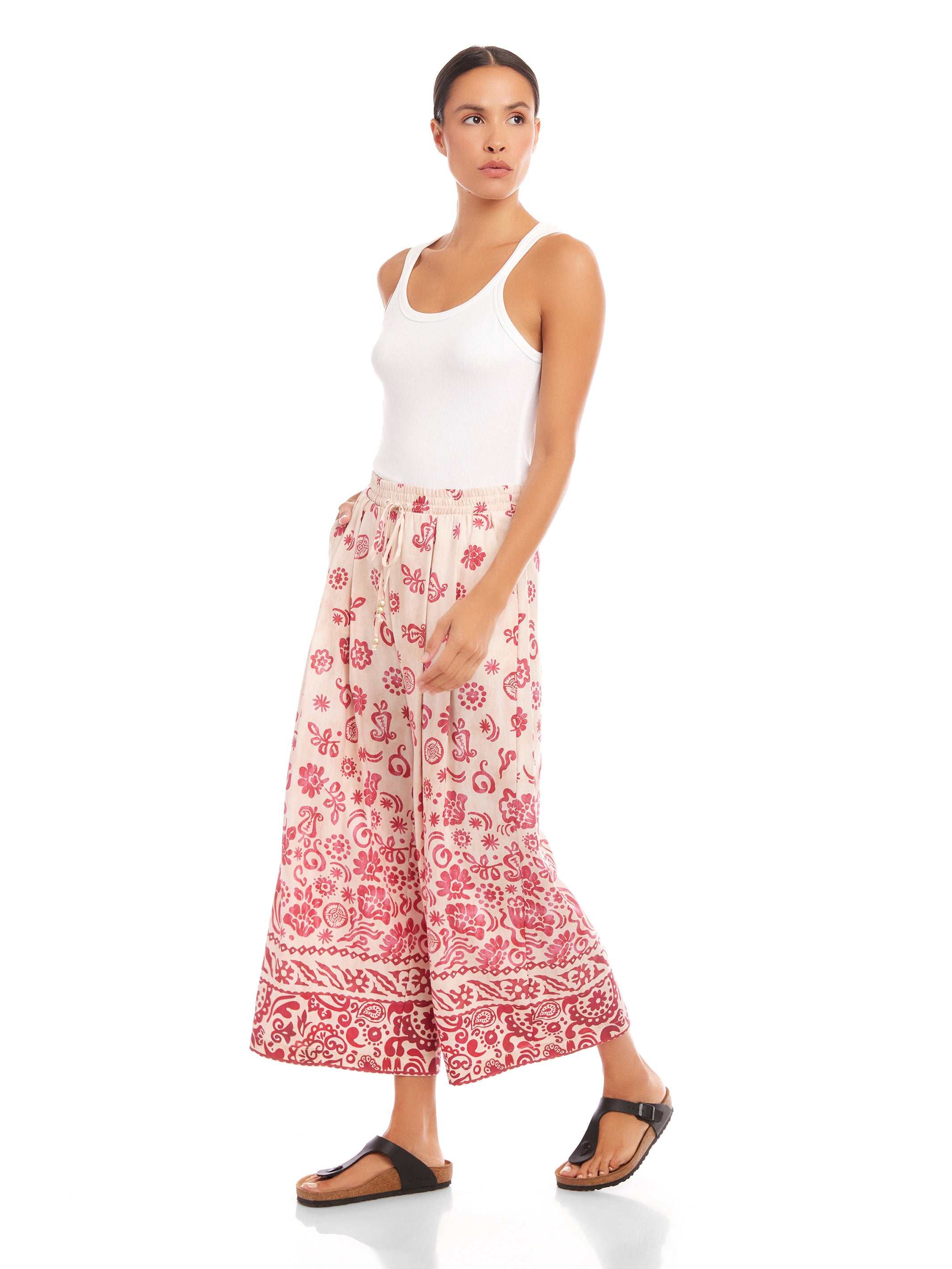Woman wearing Etta Border Print Pants with wide leg and watercolor border print in red on cream, paired with white tank top and black sandals.