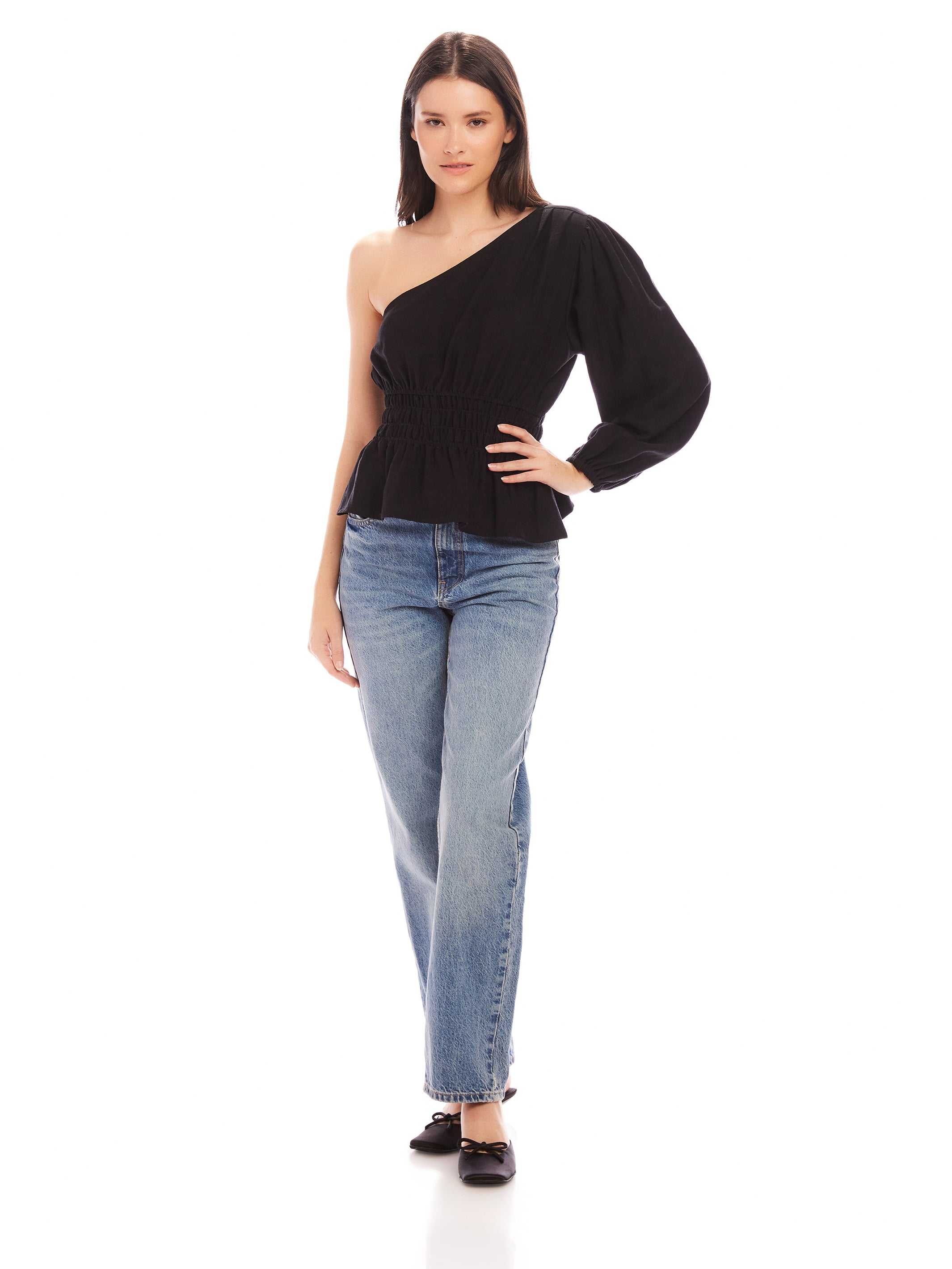 Woman wearing a black Talia One-Shoulder Top with elastic waist and asymmetrical long sleeve paired with blue jeans and black flats