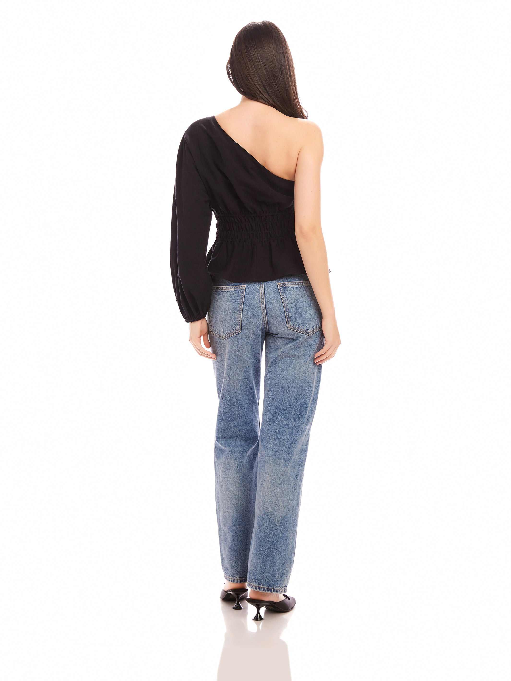 Woman wearing black Talia One-Shoulder Top with elasticized waist and asymmetrical long sleeve, paired with blue jeans.