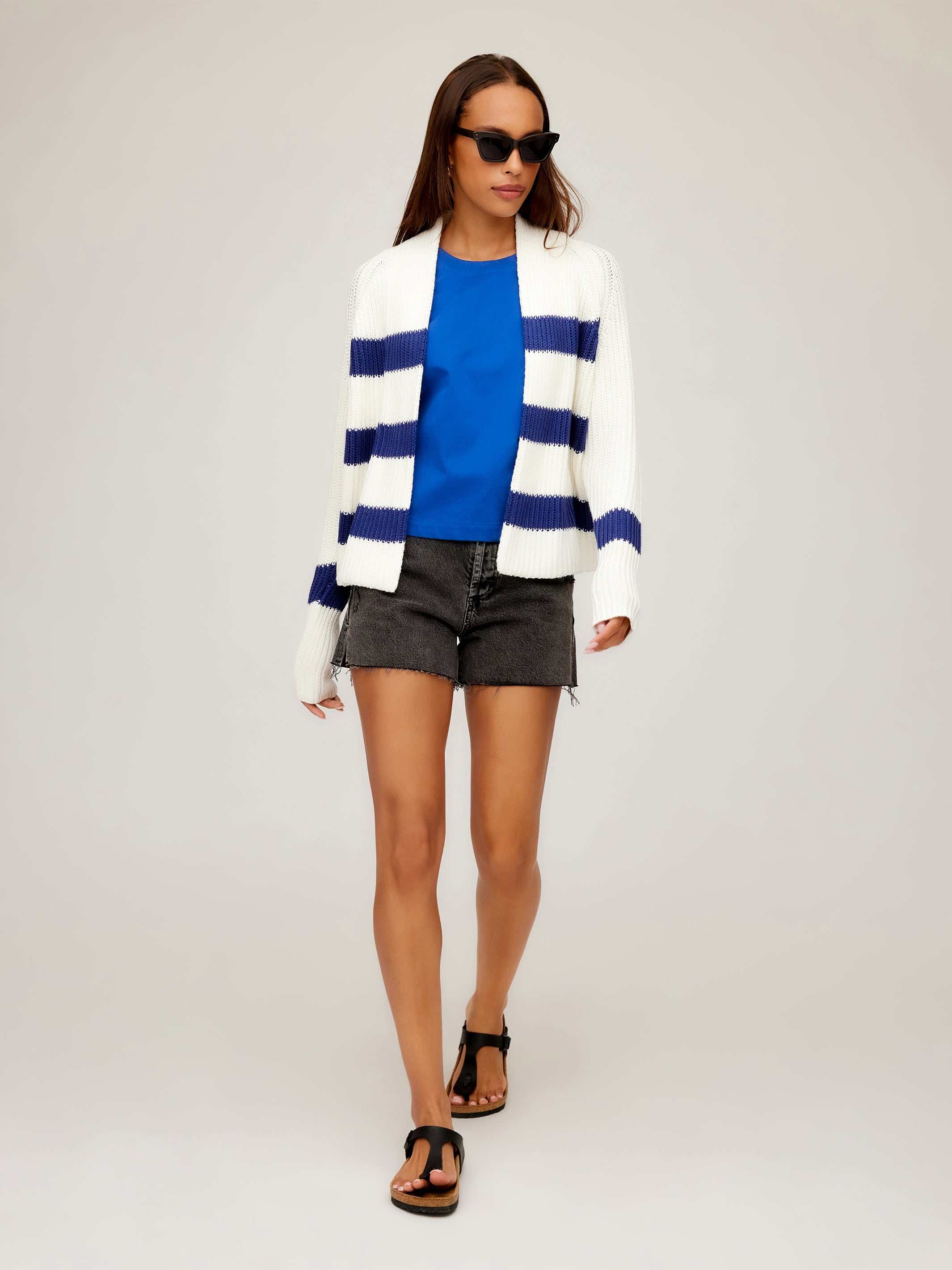 Woman wearing Cely Cropped Cardigan with white and navy stripes, blue top, black shorts, and sandals walking against plain background