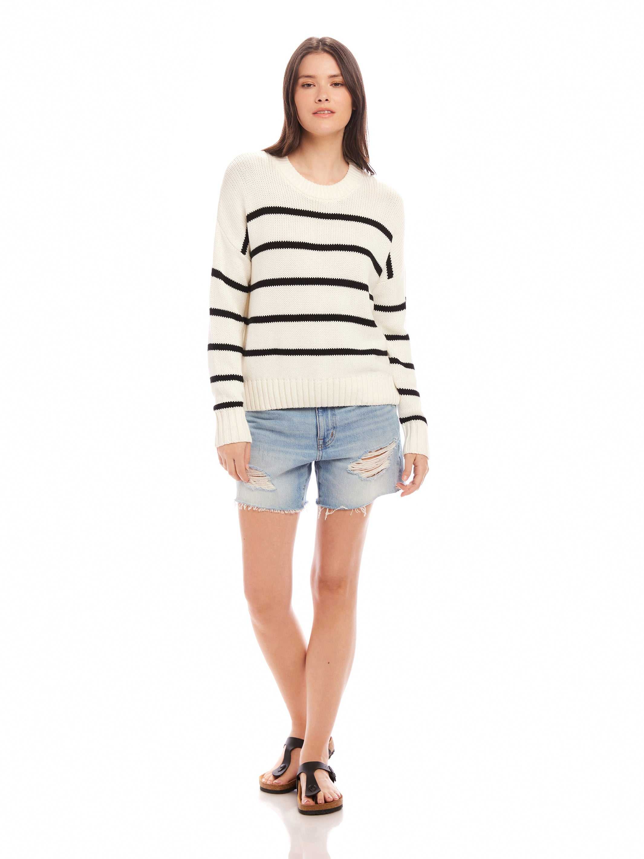 Woman wearing a white Luca Crewneck Sweater with black stripes, paired with denim shorts and sandals.