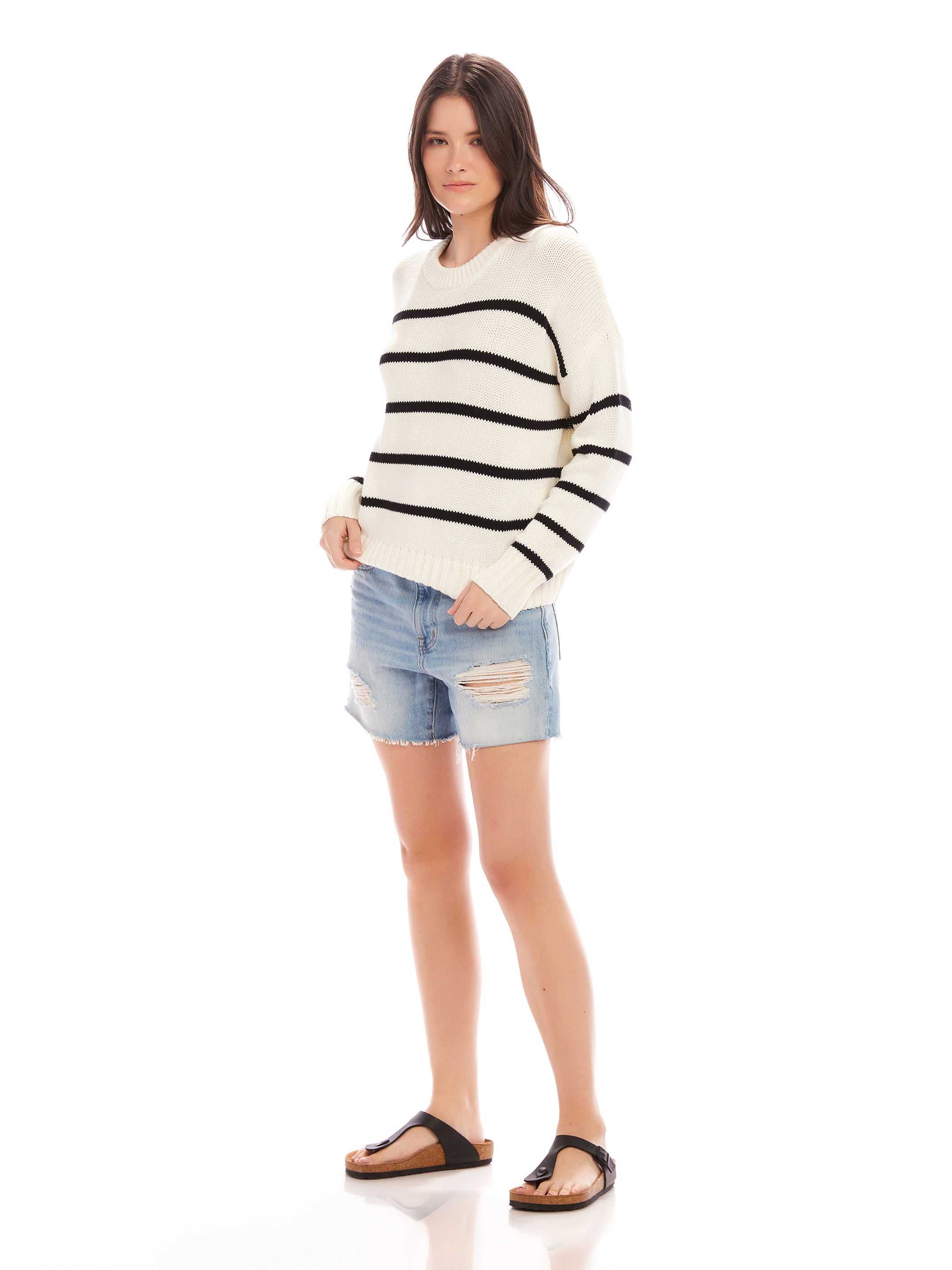 Woman wearing a Luca Crewneck Sweater with black horizontal stripes, paired with ripped denim shorts and black sandals.