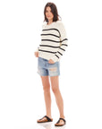 Woman wearing a Luca Crewneck Sweater with black horizontal stripes, paired with ripped denim shorts and black sandals.