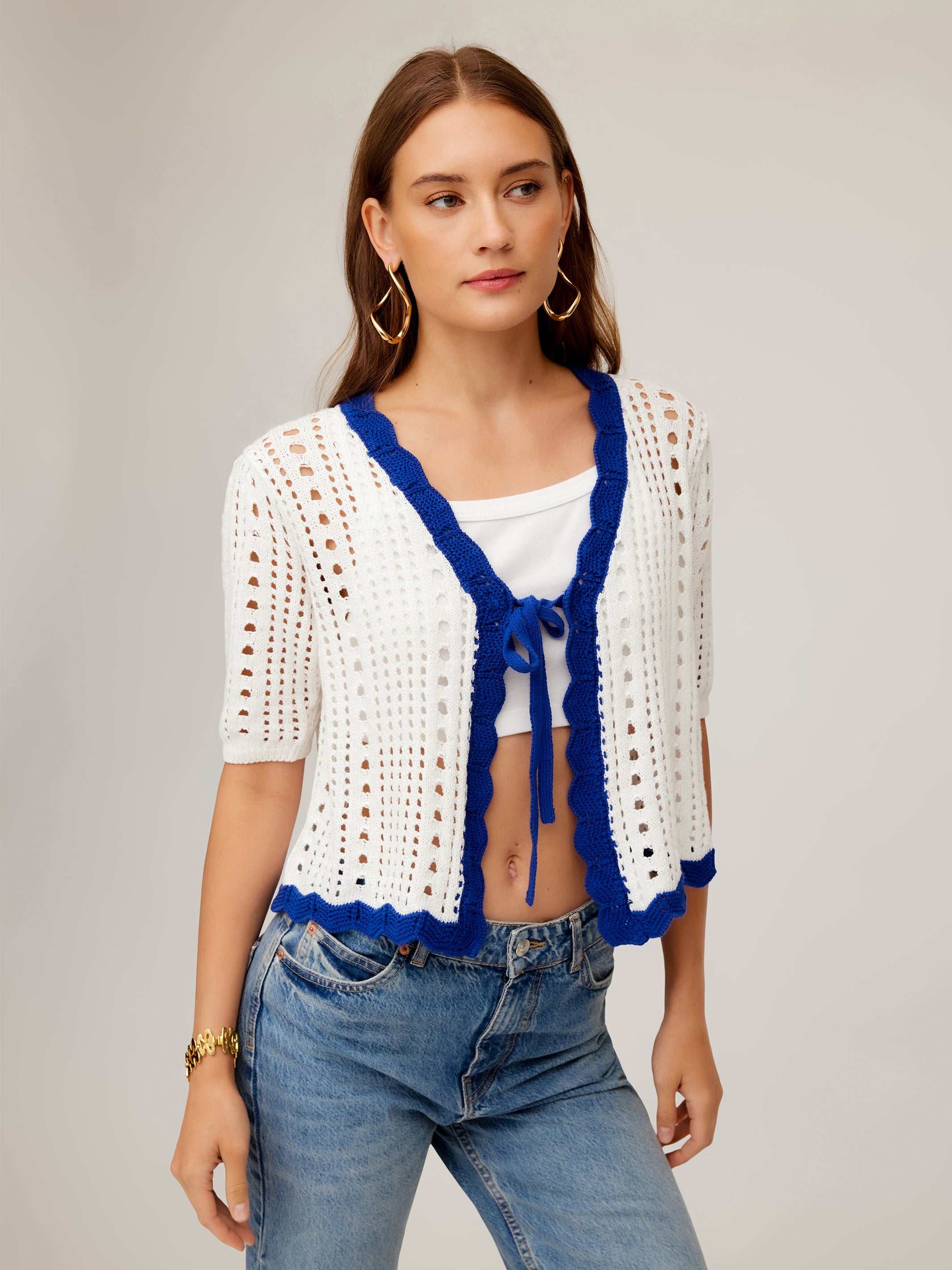 Woman wearing Betsy Crochet Top with white crochet eyelet stitching, blue scalloped trim, tie front, short sleeves, and cropped fit