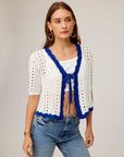 Woman wearing Betsy Crochet Top with white crochet eyelet stitching, blue scalloped trim, tie front, short sleeves, and cropped fit