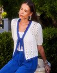 Woman wearing Betsy Crochet Top with white crochet eyelet stitching and blue scalloped trim, styled open with tie front and short sleeves