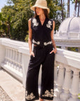 Woman wearing black Mason Rope Stitch pants and matching sleeveless vest with cream embroidery, standing outdoors by a white railing and palm trees.