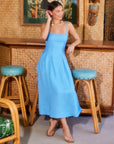 Woman wearing sleeveless blue Jessa Midi Dress with adjustable straps and side pockets in tropical-themed room