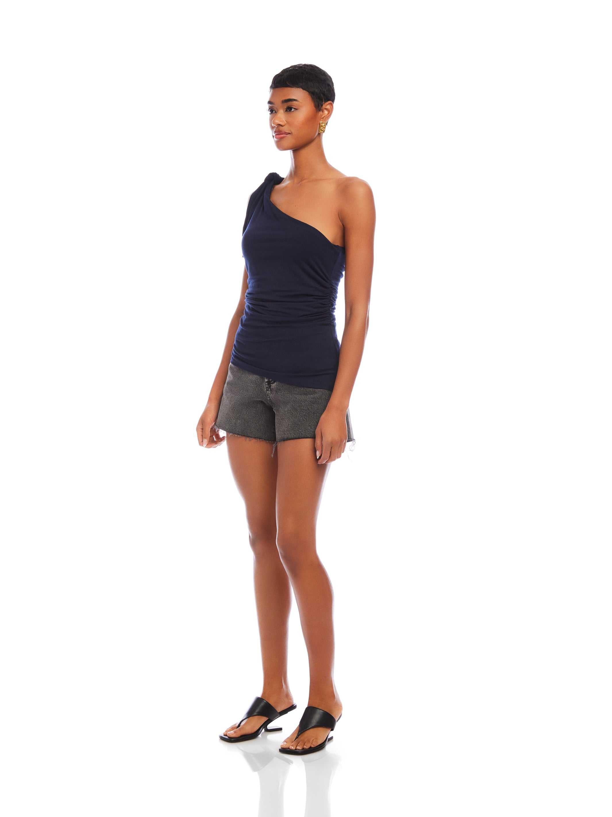 Woman wearing navy Harper One-Shoulder Tie-Top with tie detail and asymmetrical silhouette paired with black shorts and sandals