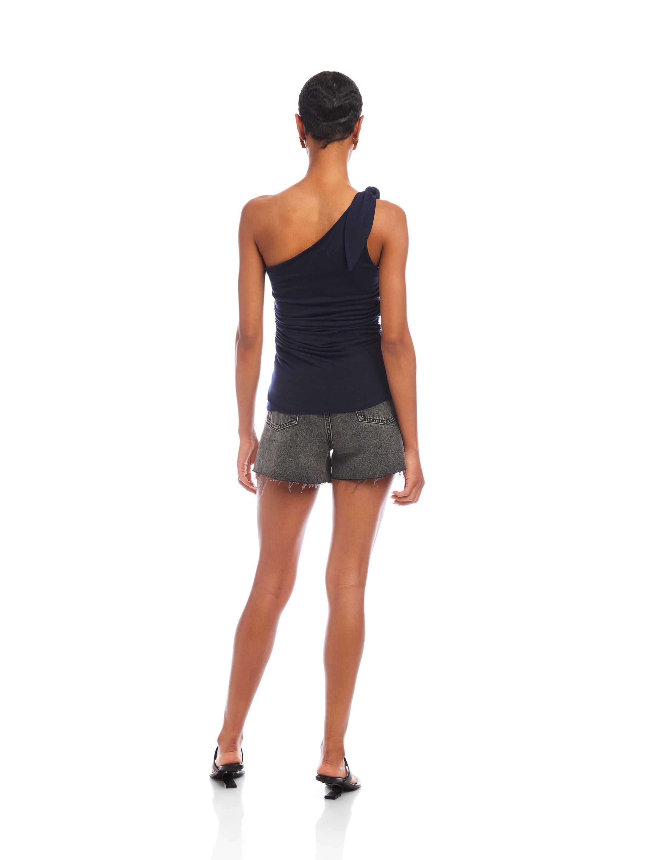 Woman wearing navy Harper One-Shoulder Tie-Top with sleeveless asymmetrical silhouette and tie detail on shoulder paired with gray shorts