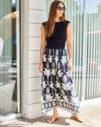 Woman wearing Carina Long Contrast Dress with sleeveless black fitted top and flowing skirt with white palm print, standing outdoors with sunglasses.